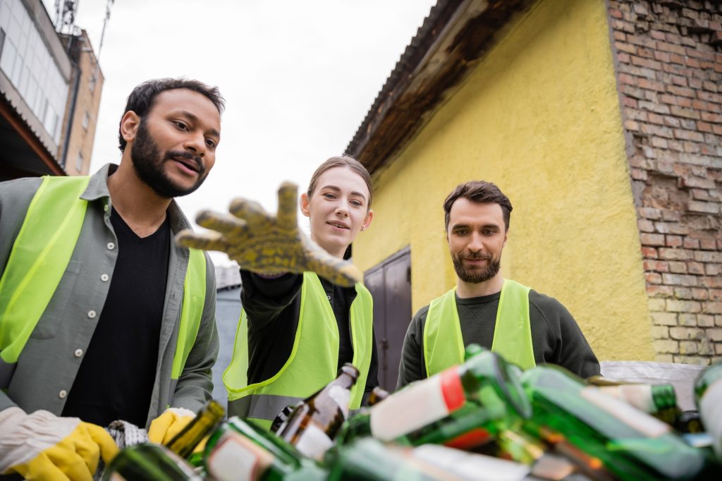 Smiling multiethnic workers in gloves and protective vests looking at blurred glass trash